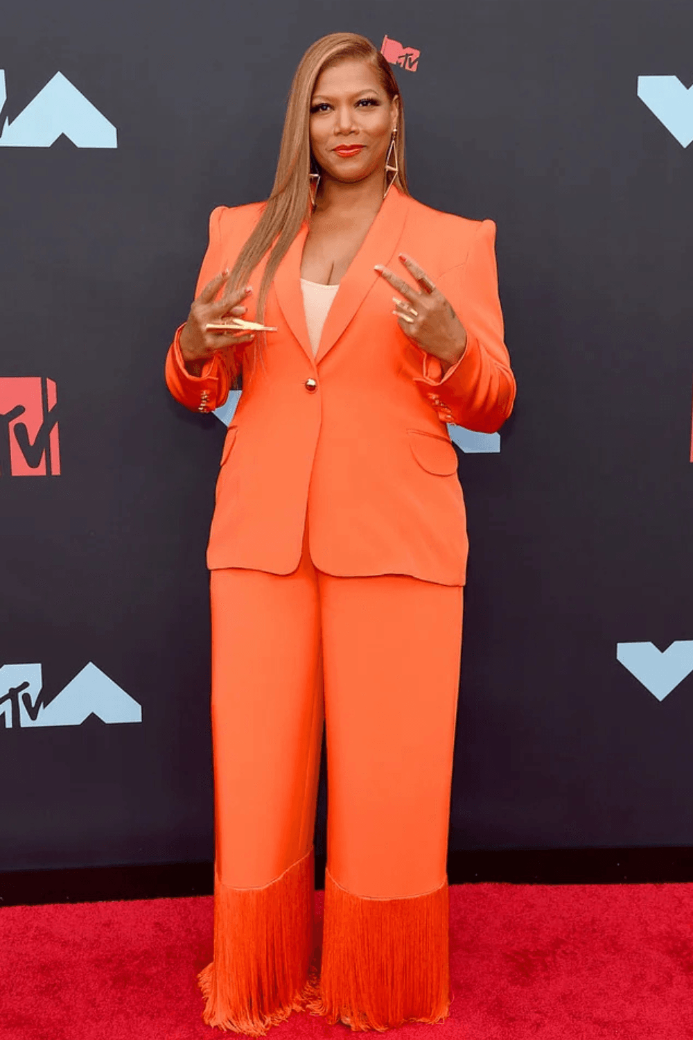 Persona de pie en una alfombra roja delante de un tel&oacute;n de fondo de la MTV, con un traje naranja con largos flecos en los dobladillos del pantal&oacute;n, mostrando c&oacute;mo vestir si tienes una silueta ovalada, posando con confianza con ambas manos levantadas cerca del pecho.