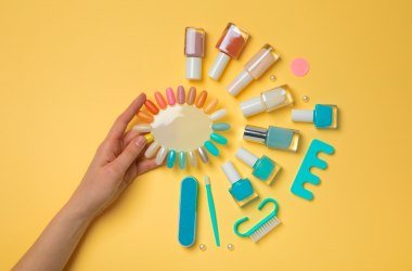 Una mano sosteniendo una paleta de vibrantes colores de uñas, rodeada de varios esmaltes de uñas, herramientas de manicura y pinceles sobre un fondo amarillo.