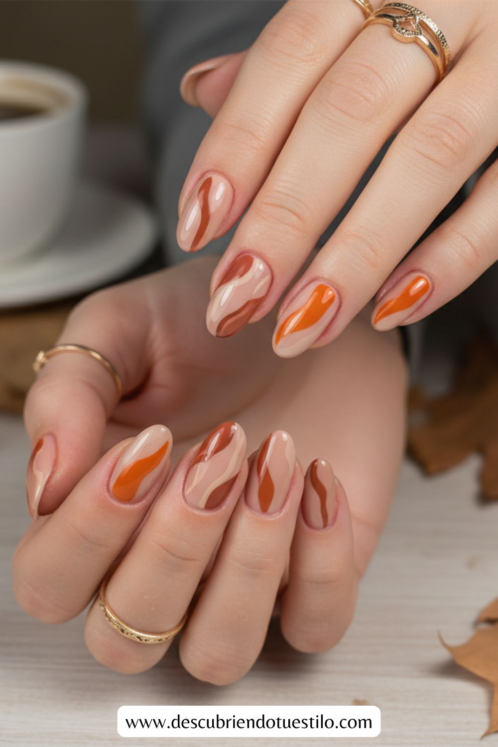 Manos con uñas largas y almendradas que presentan un diseño abstracto y ondulado en beige, naranja y marrón: perfectas uñas otoñales. Varios anillos dorados adornan los dedos para un look elegante y festivo ideal como uñas para Acción de Gracias.