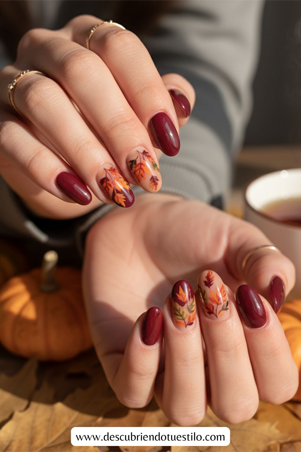 Primer plano de unas manos con uñas elegantes y decoración de uñas con motivos de hojas de otoño, sosteniendo una taza de té; en el fondo se ven pequeñas calabazas, perfectas para uñas para Acción de Gracias.