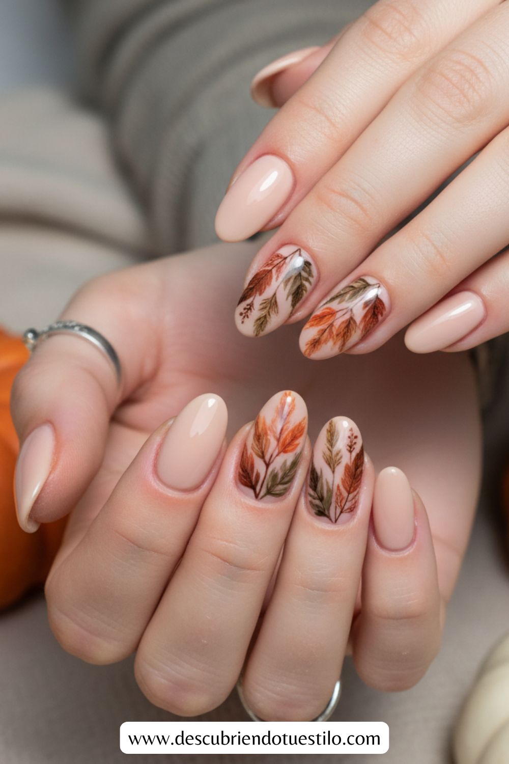 Primer plano de manos con uñas elegantes en forma almendrada pintadas nude; dos uñas en cada mano lucen diseños otoñales de hojas en tonos naranjas, marrones y verdes, perfectas como uñas para Acción de Gracias.