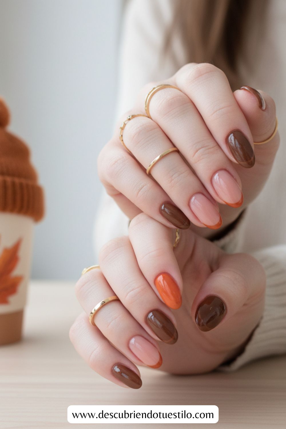 A person displays uñas otoñales pintadas en tonos marrón, nude y naranja mientras luce anillos dorados. Una taza con diseño de hoja adorna la mesa al fondo. Ideal inspiración para uñas para Acción de Gracias.