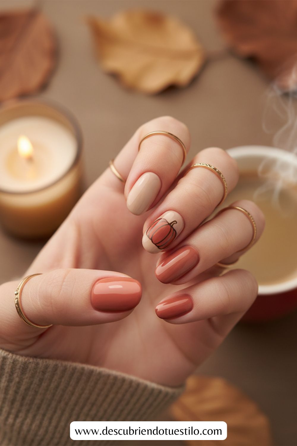 Una mano con uñas otoñales en naranja, nude y calabaza, con anillos dorados, una vela encendida, una taza de café y hojas caídas de fondo: uñas perfectas para Acción de Gracias.