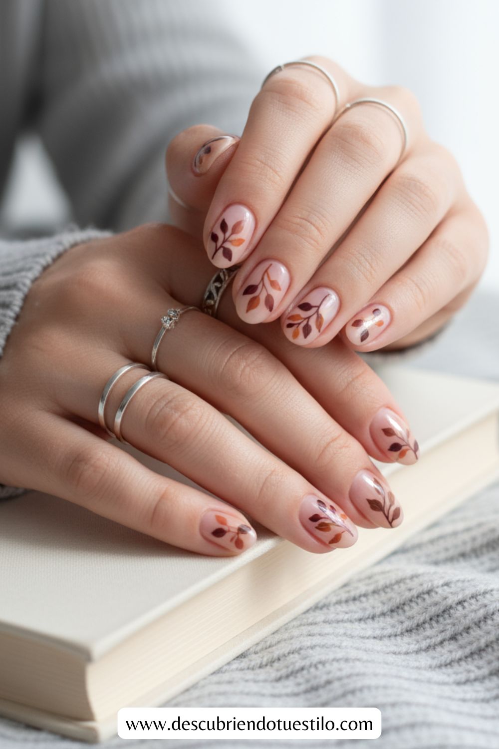 Primer plano de unas manos con uñas otoñales con motivos de hojas en color nude y marrón, apoyadas en un libro cerrado. La persona lleva anillos de plata en varios dedos.